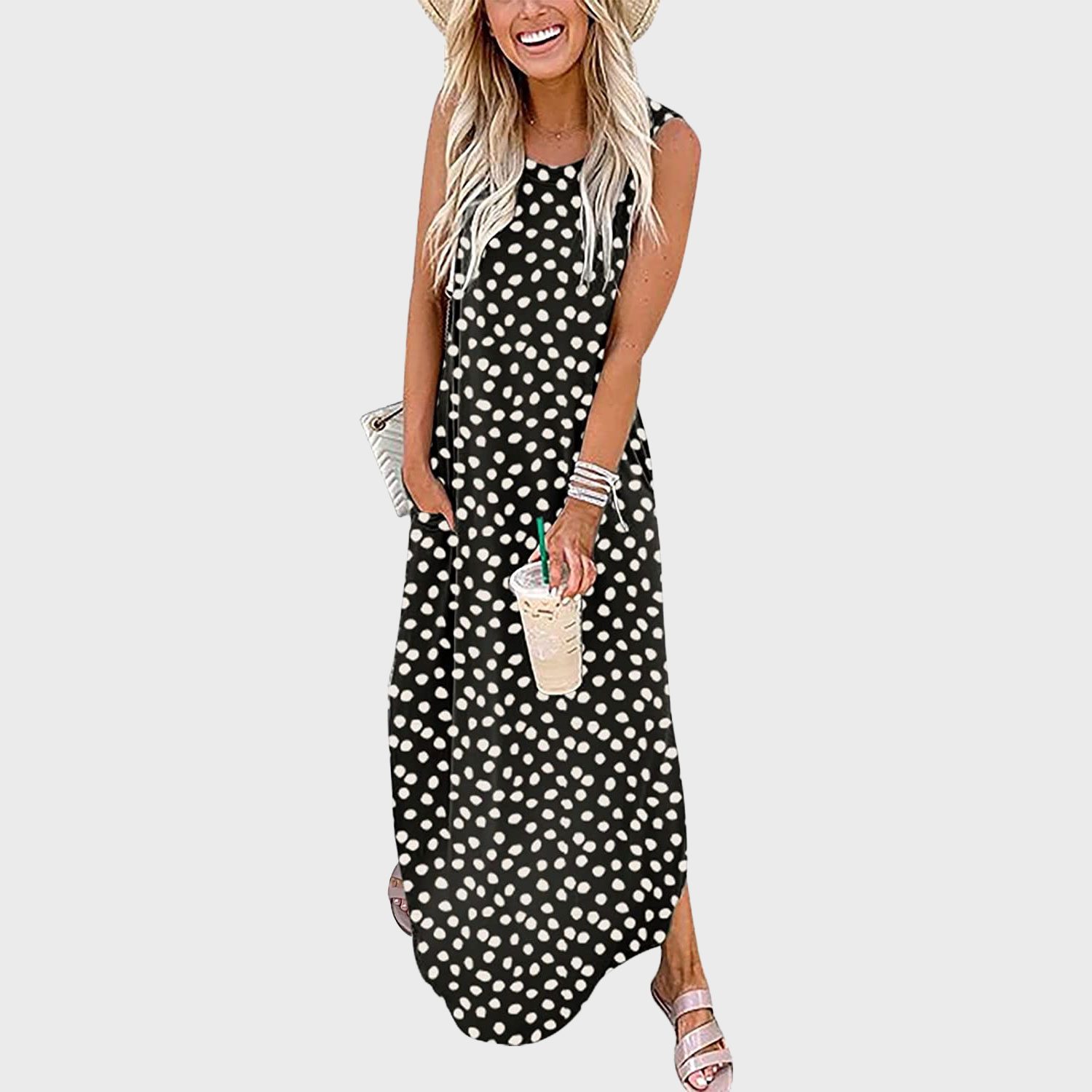 A woman in a polka-dot dress holds a drink and smiles, wearing a hat, bracelets, and sandals, on a plain background.