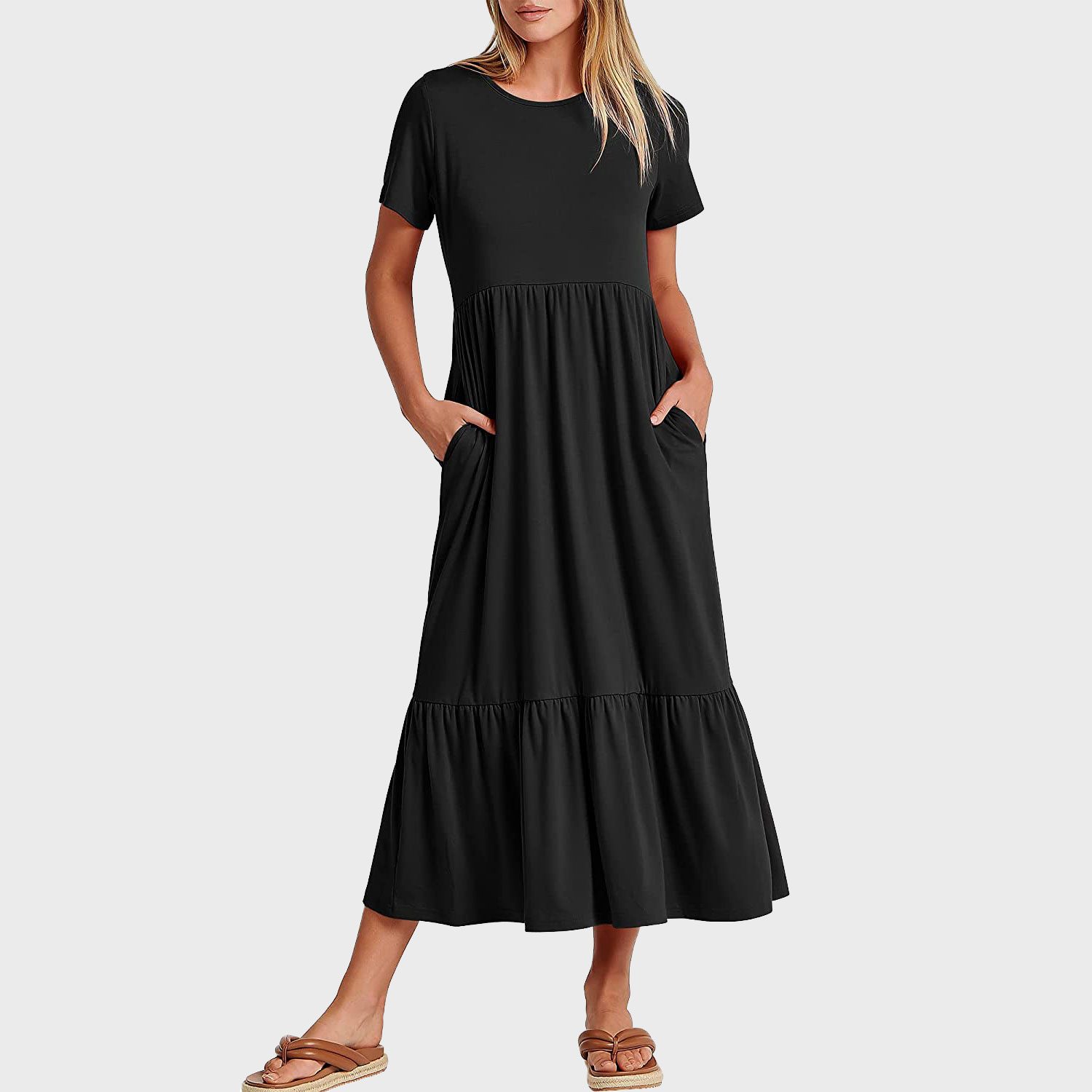 A woman stands wearing a black maxi dress with short sleeves and tiered skirt, hands in pockets, against a gray background.