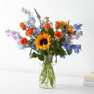 Vase holds colorful flowers, including sunflowers, roses, and blue blooms, placed on a white surface beside stacked books.