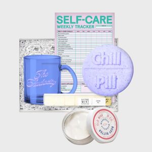 Blue mug and purple bath bomb rest on a self-care weekly tracker, surrounded by luxury soap and body butter, all in a gift box.