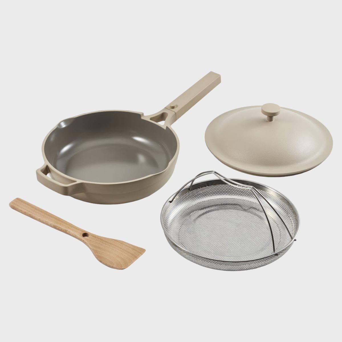Cookware set includes a beige frying pan, matching lid, metal steamer basket, and wooden spatula on a plain white background.