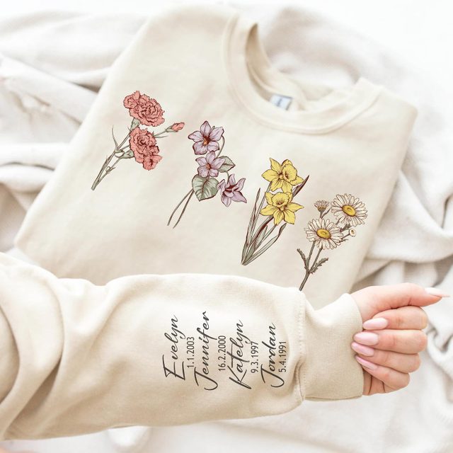 Beige sweatshirt displays colorful floral illustrations with handwritten names and dates on the sleeve, held by a hand with manicured nails in a bright setting.