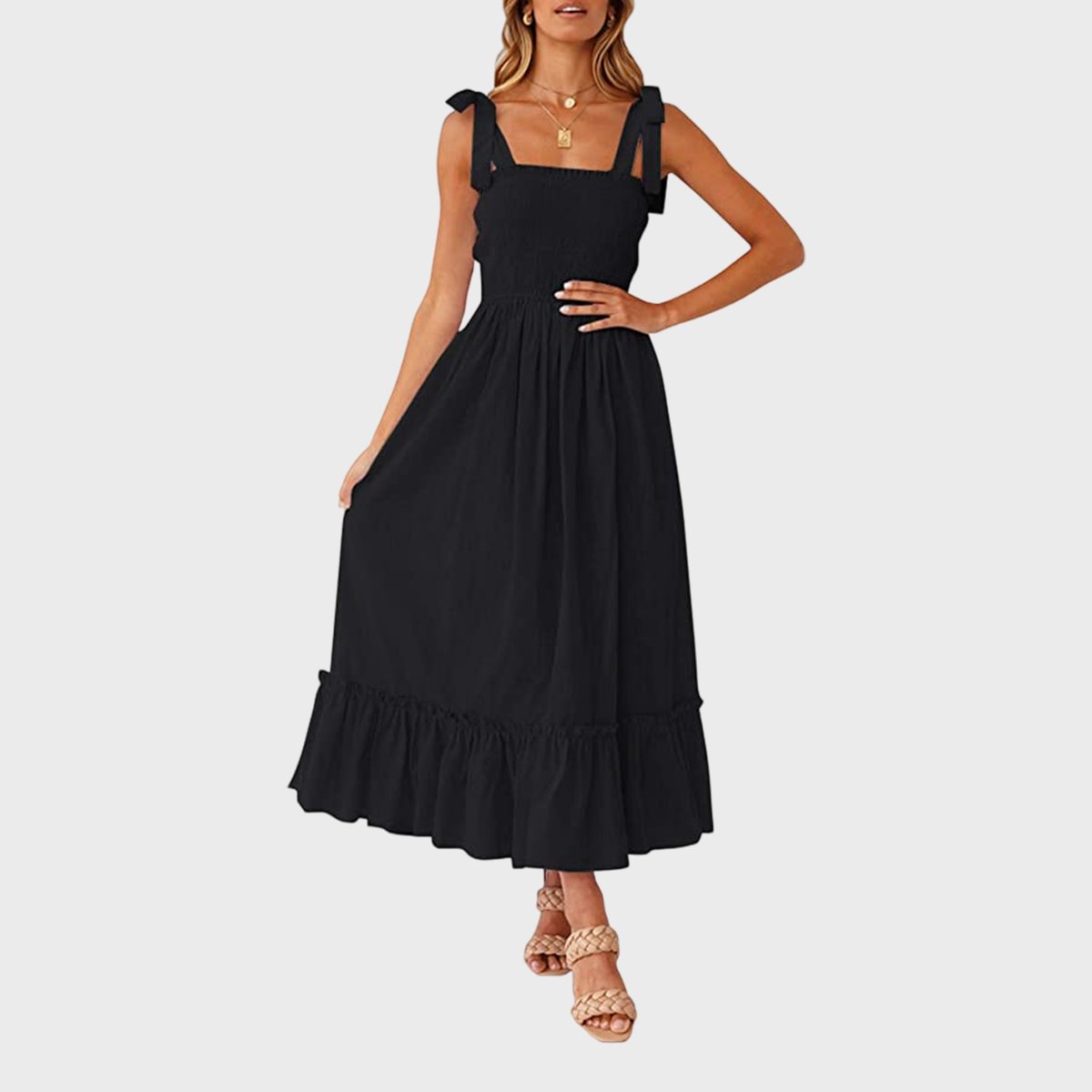 A woman poses in a sleeveless black dress with bows, wearing tan braided sandals against a plain white background.