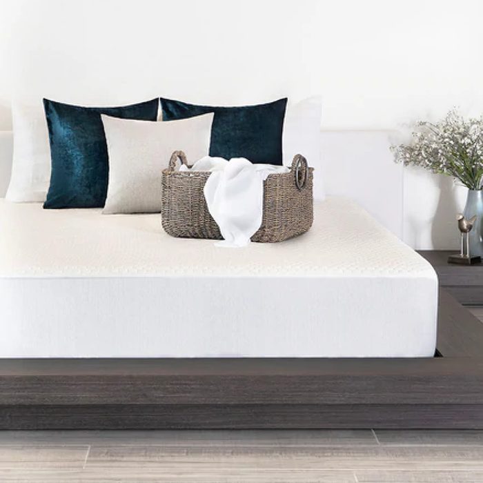 Basket holds folded white towel on a neatly made bed with pillows in a modern bedroom, decorated with flowers in a vase.