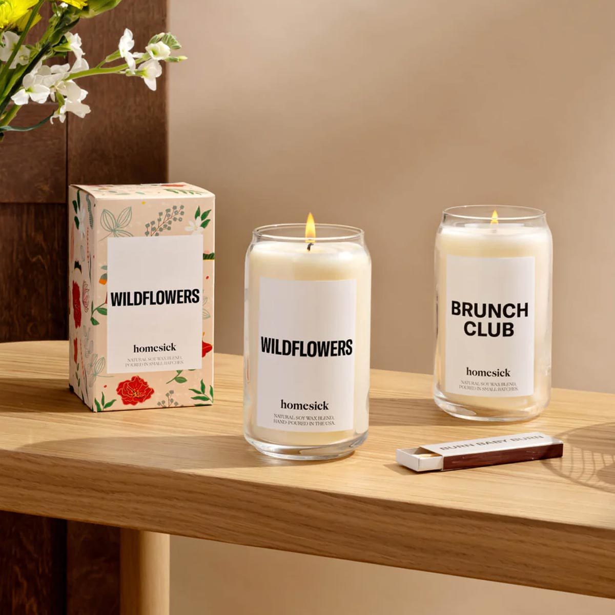 Candles burn on a wooden table, labeled "WILDFLOWERS" and "BRUNCH CLUB." A box and matchbox are nearby; flowers are visible in the background.