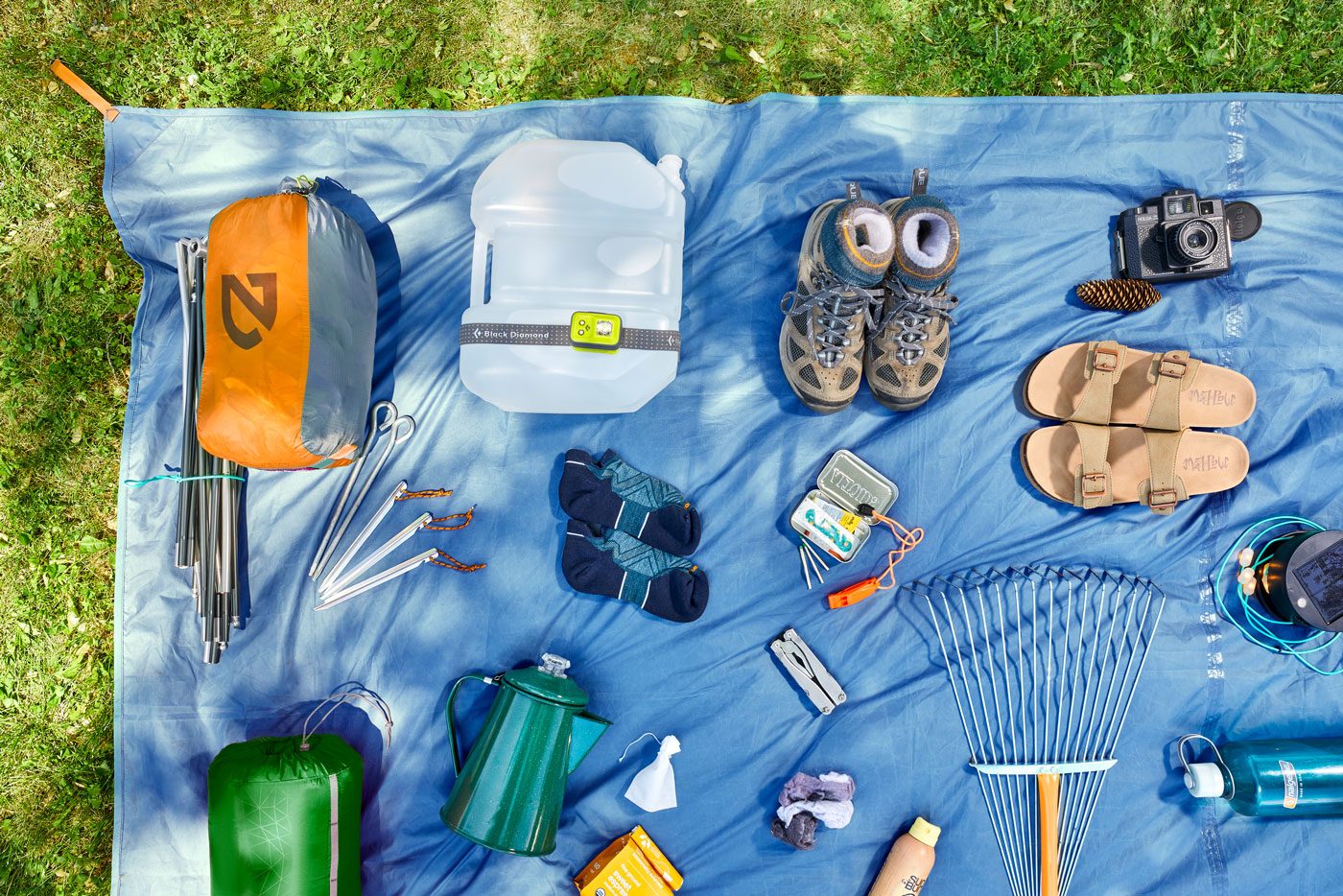 Camping gear arranged on a blue tarp, surrounded by grass; includes backpacking tent, water jug, hiking boots, sandals, camera, utensils, and safety equipment.