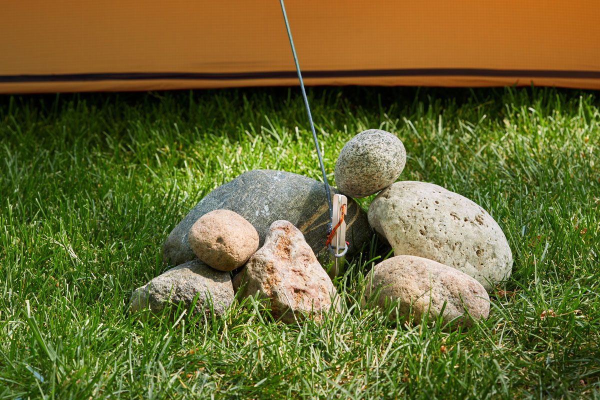 Tent stake secures orange fabric, surrounded by rocks on grassy ground.