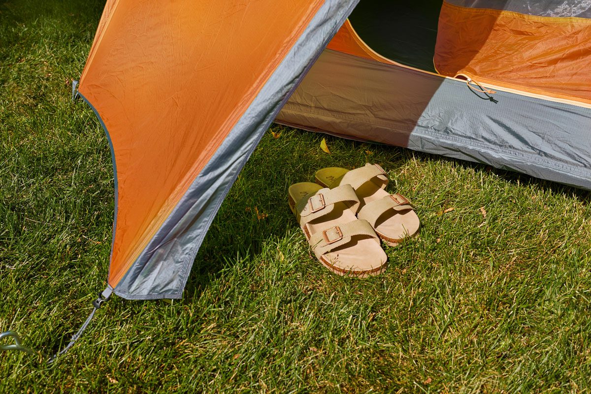 Sandals resting beside an orange-gray tent entrance on a grassy field.
