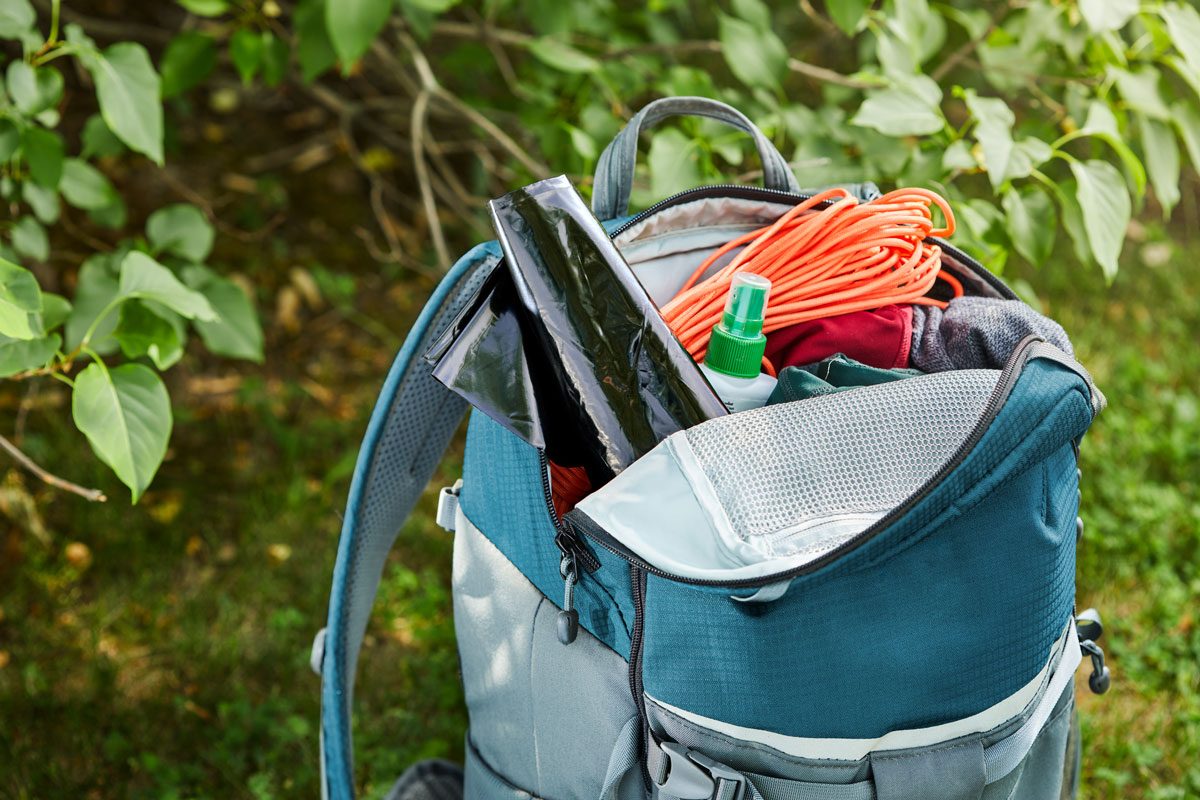 Backpack holding gear: rope, bottle, and bags, situated outdoors among green leaves and grass.