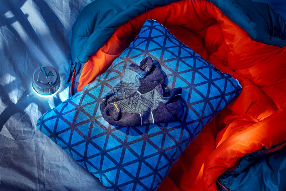 Socks rest on a patterned blue pillow beside an opened blue and orange sleeping bag on a bed, with a small light nearby.