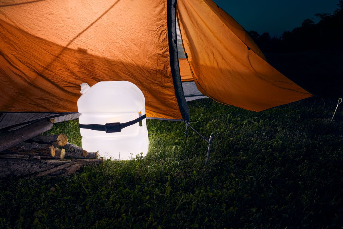 A translucent water container glows beside chopped firewood near an orange camping tent on grassy ground at night.