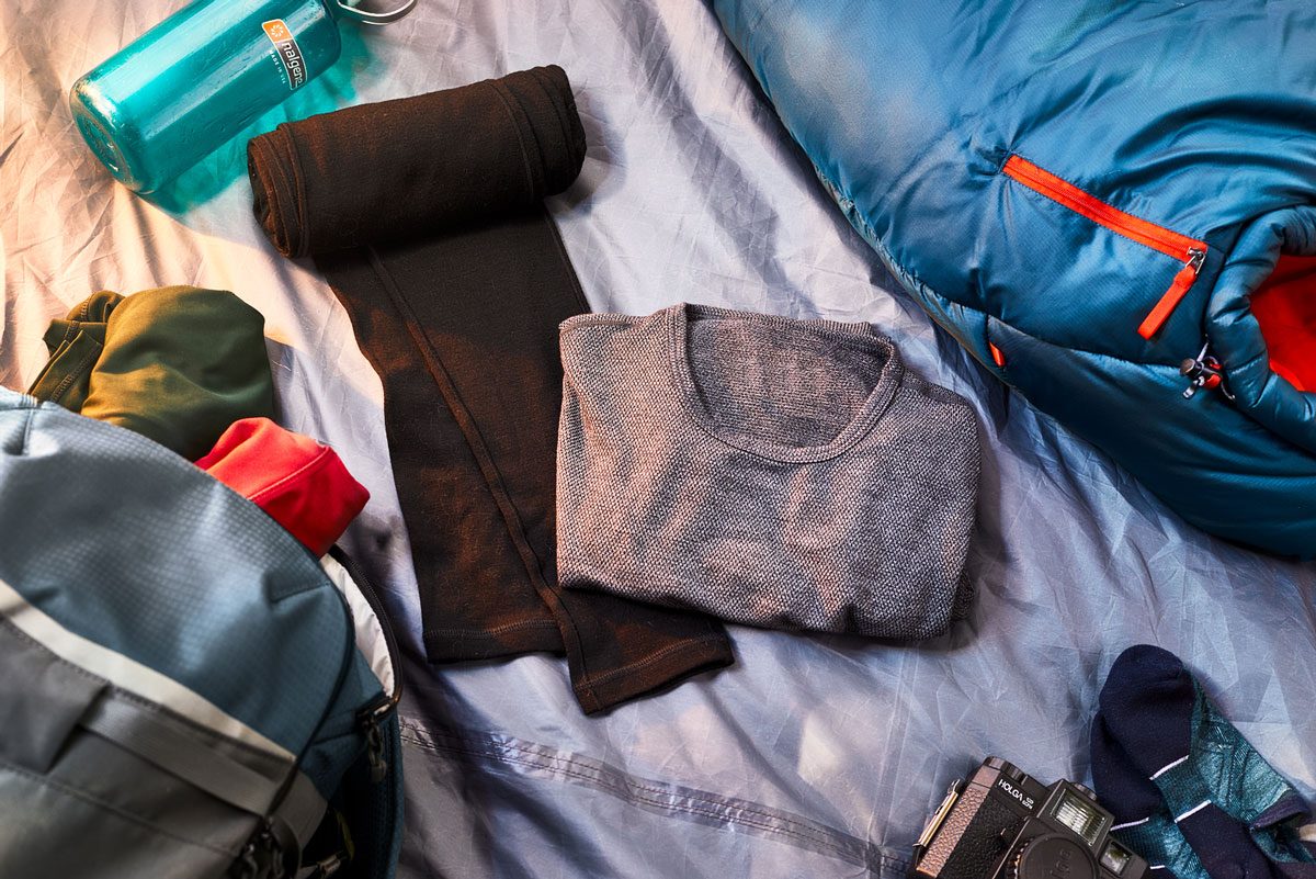Clothing and gear arranged; a shirt, pants, sleeping bag, camera, and a water bottle lie on a fabric surface, surrounded by a backpack and socks.