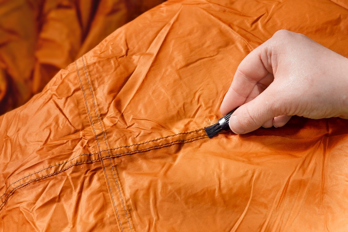 A hand applies sealant over orange fabric seams using a small brush, suggesting repair or waterproofing in a domestic setting.