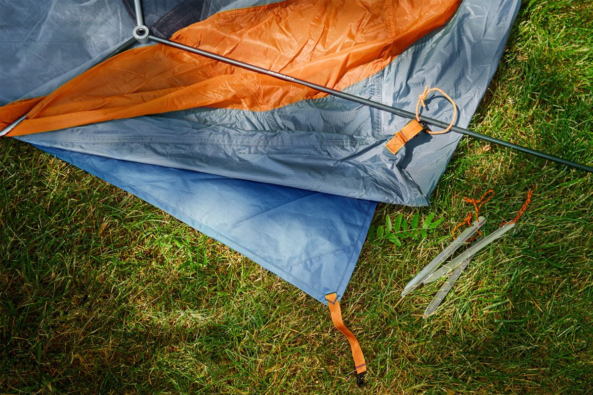 A tent setup in progress, featuring colorful fabric and poles, laid out on green grass with tent stakes nearby.