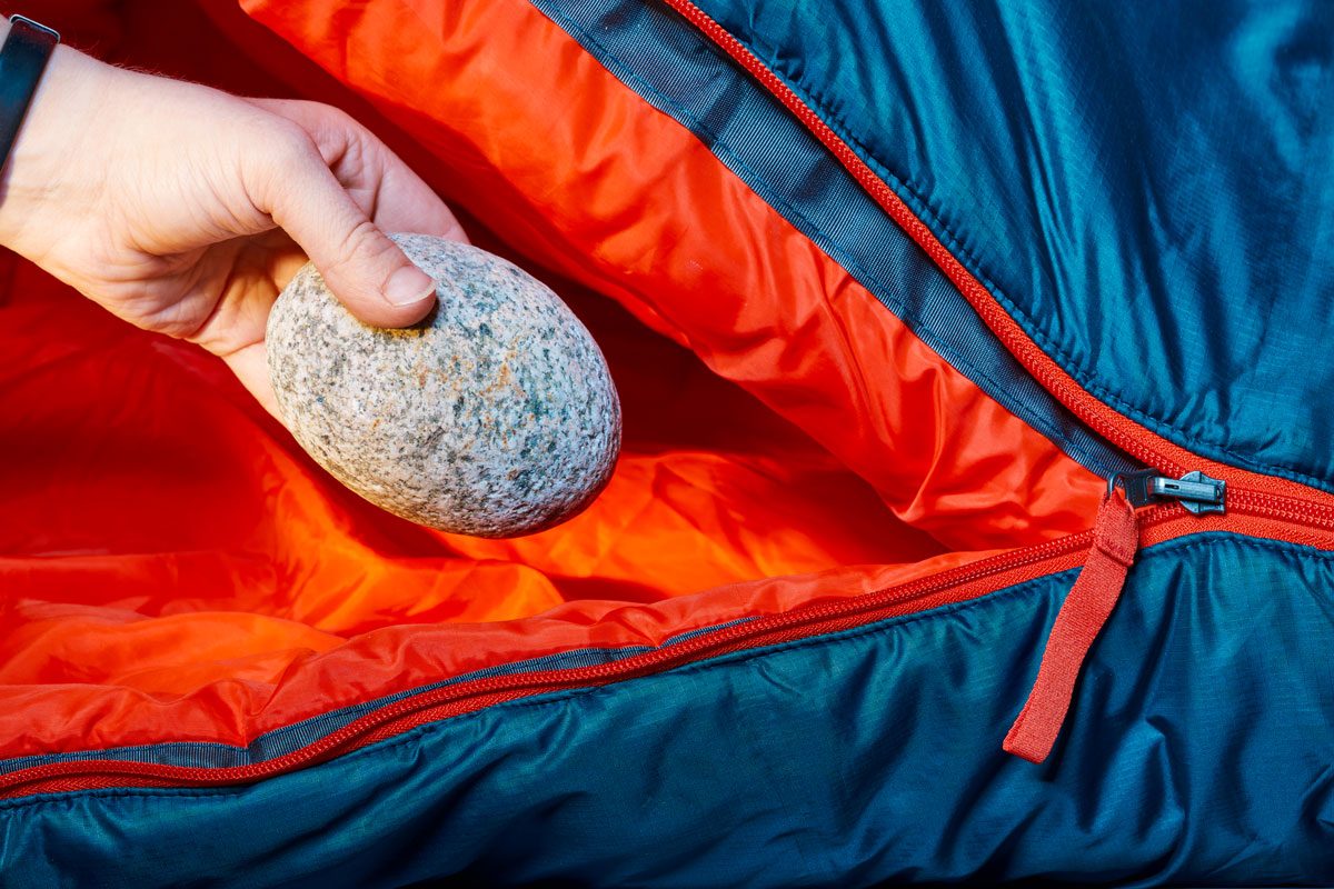 A hand places a stone inside a partially unzipped red and blue sleeping bag.