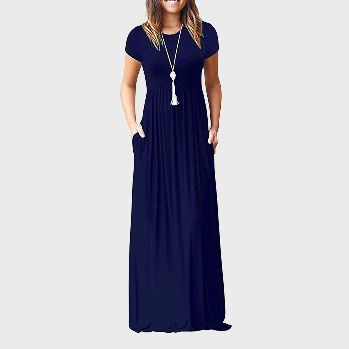 A woman wearing a long, navy blue dress with pockets and a tassel necklace, standing against a light gray background.