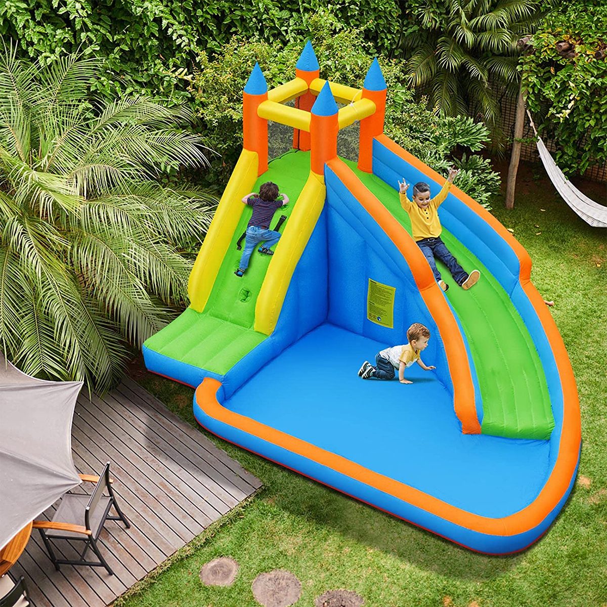 Inflatable bouncy castle with two slides; children playing energetically; sunny backyard with lush greenery and a patio area.