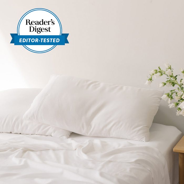 White pillows rest on a bed with rumpled sheets, next to a wooden nightstand holding white flowers. A 