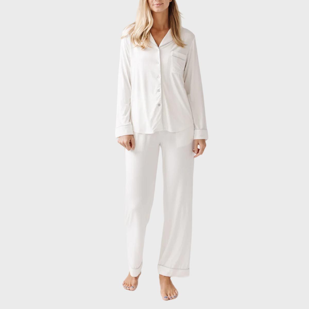 Person standing, wearing a cream pajama set with buttoned shirt and pants, barefoot, against a plain light gray background.