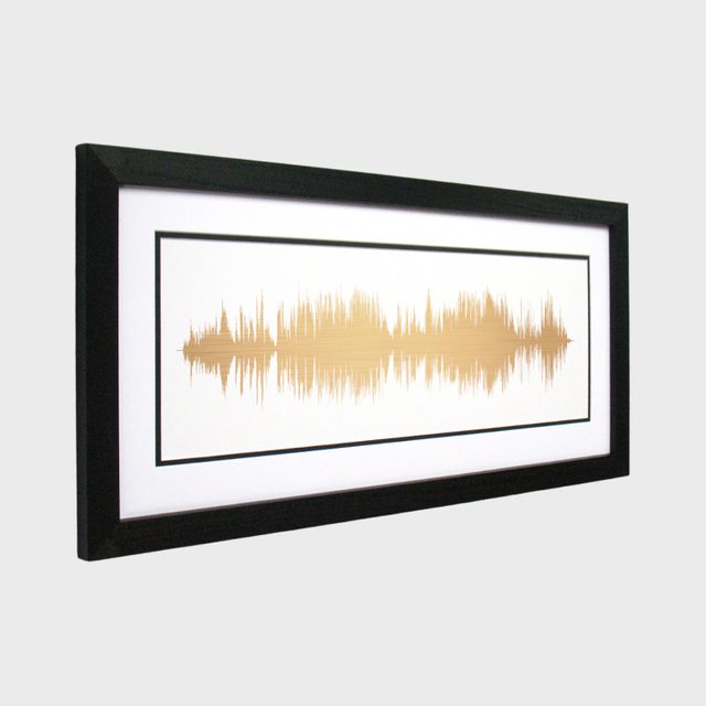 A framed artwork displays gold soundwaves on a white background, with a black border, set against a plain gray wall.
