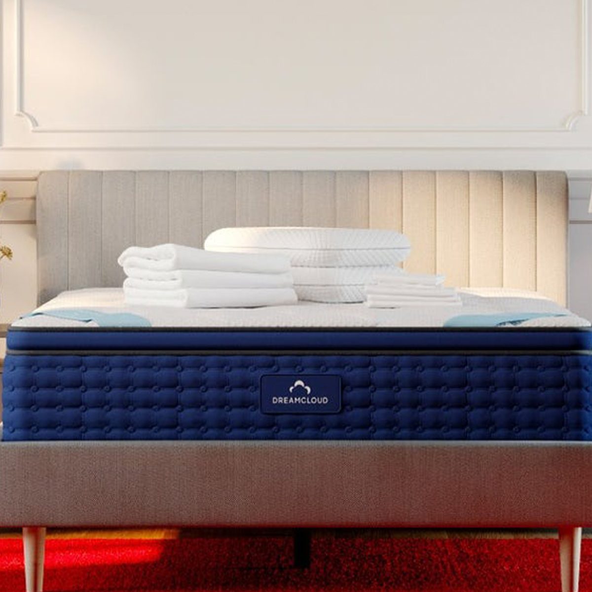Mattress stacked with folded towels sits on a bed frame in a softly lit bedroom. A navy label reads "Dreamcloud."