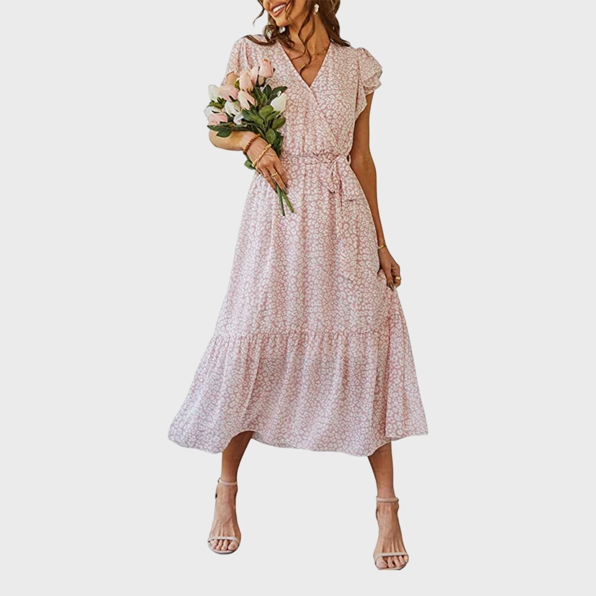 A woman holds pink flowers, wearing a light pink floral dress in a minimal, plain setting.