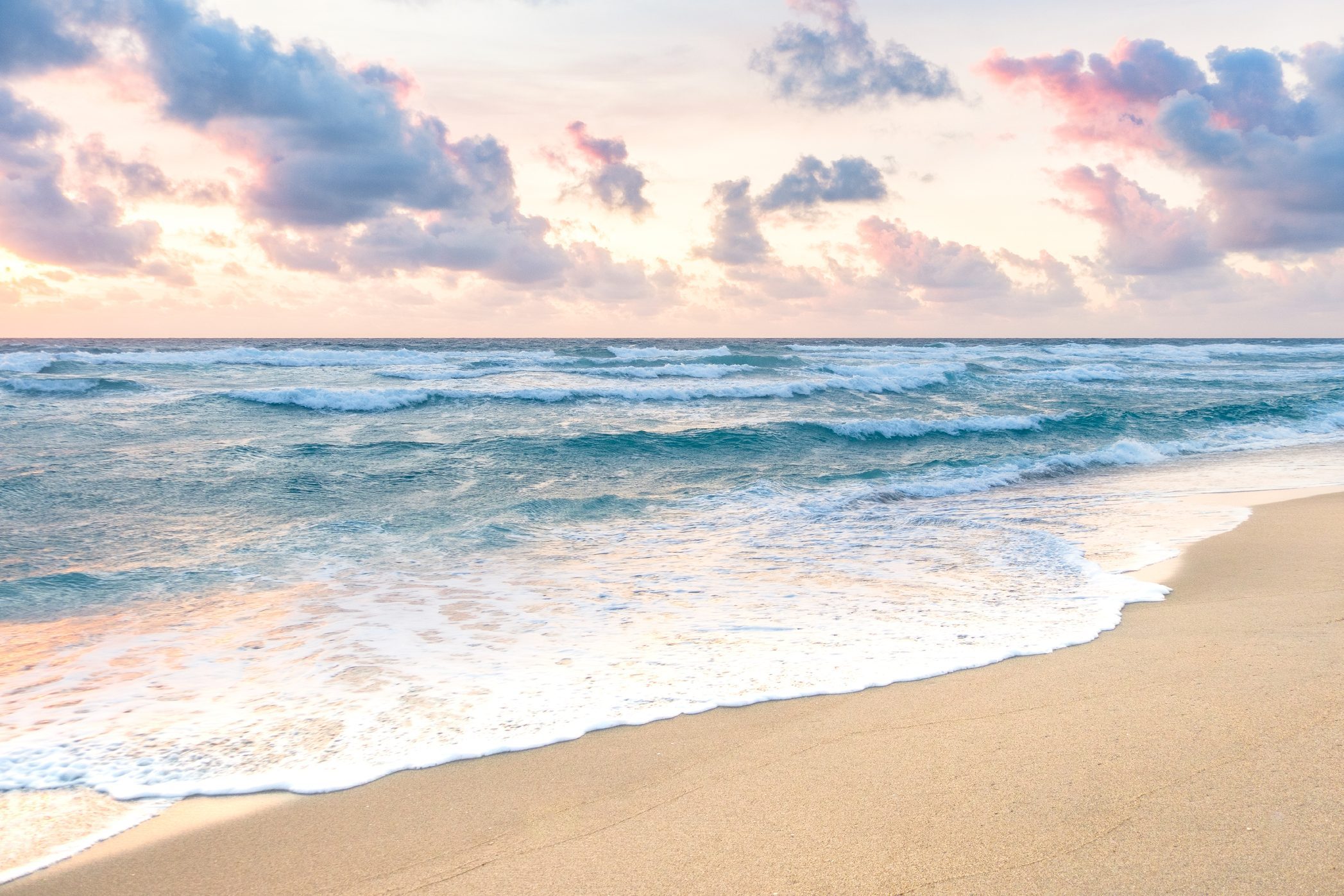 Waves gently crash on sandy beach under a sky filled with fluffy, pink-tinged clouds during sunset.
