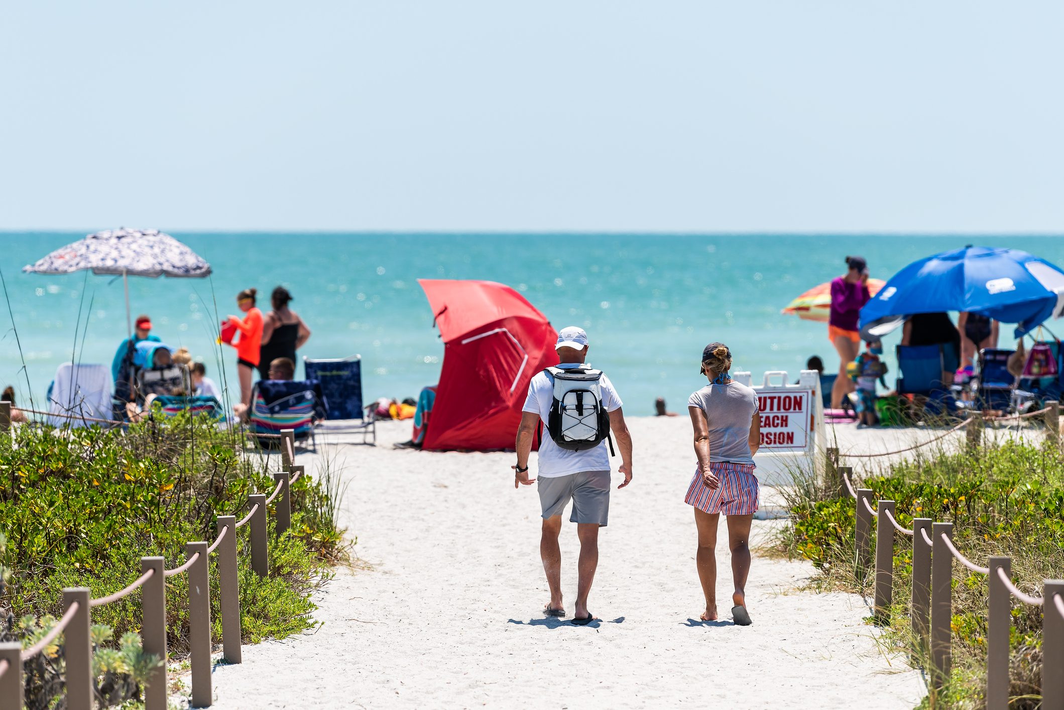 Two people walk along a sandy path towards a busy beach with umbrellas and people. A sign reads "Caution Beach Erosion."