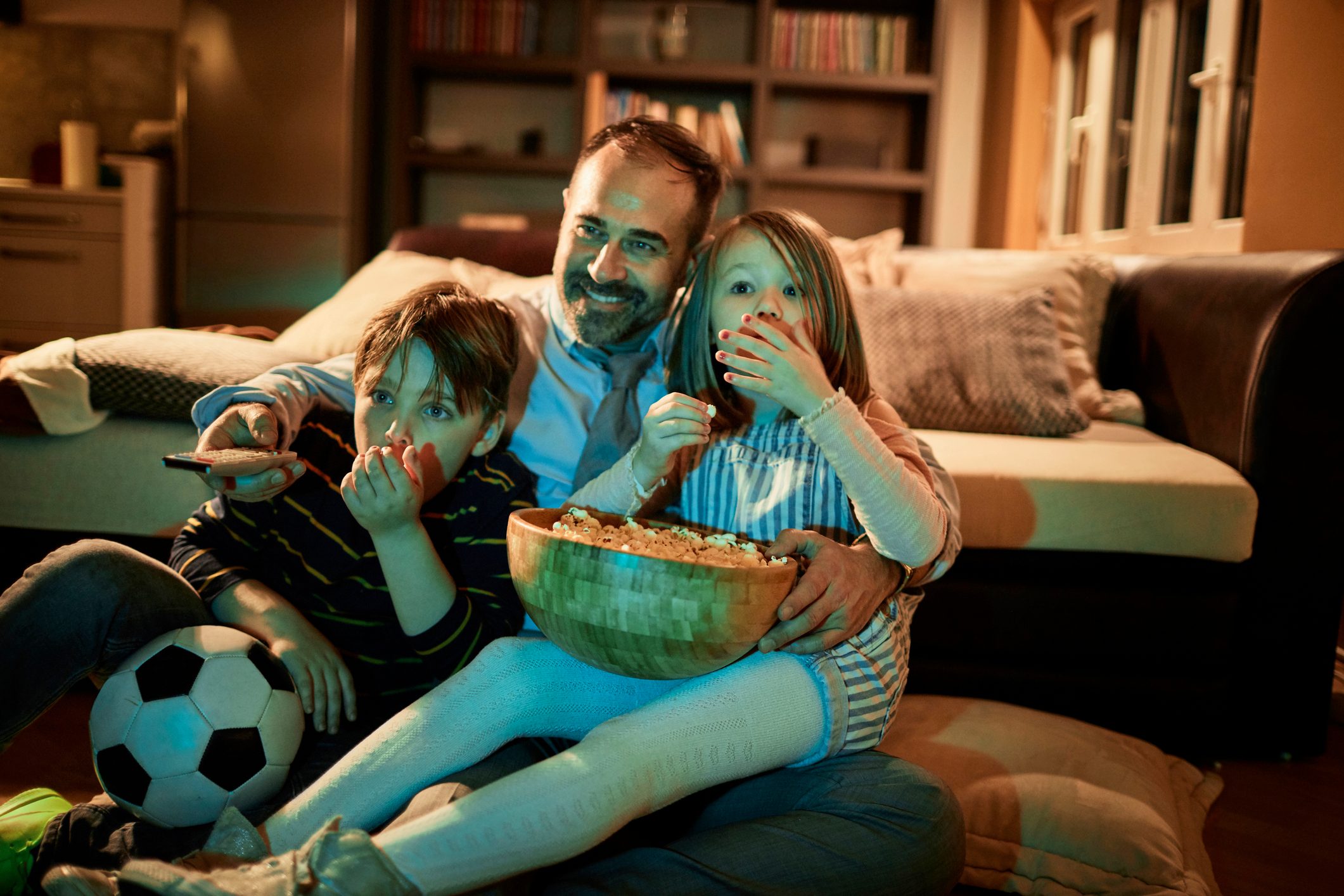 A family sits on the floor watching television, eating popcorn, and surrounding a cozy living room with bookshelves and a couch.