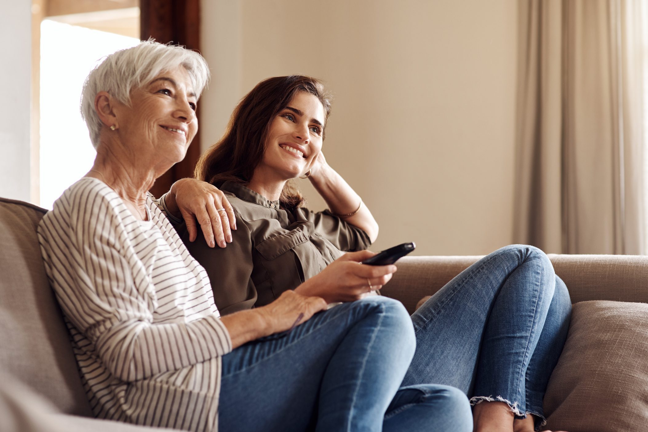 Two women sit on a couch, smiling and holding a remote, in a cozy living room setting with curtains in the background.