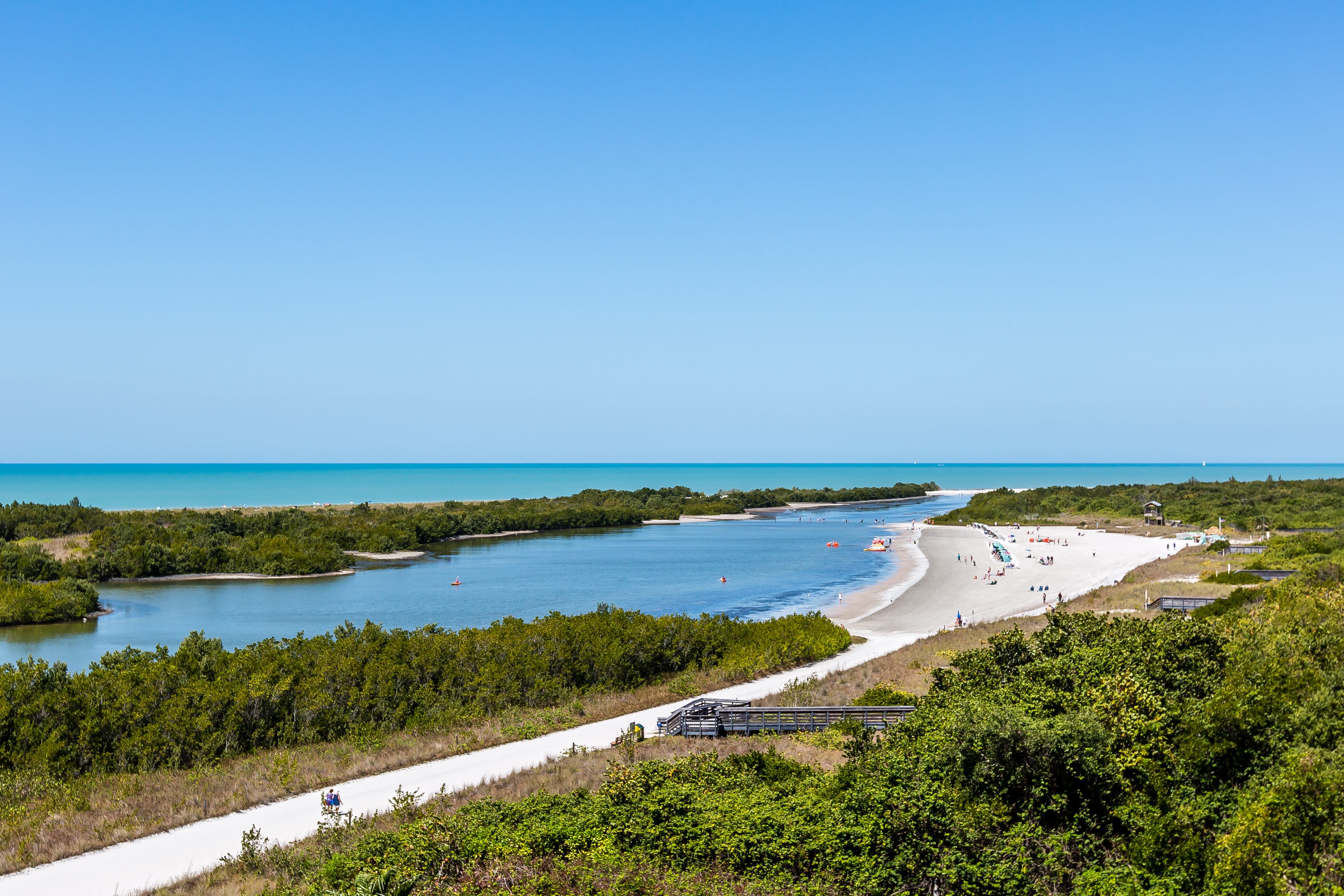 Beachgoers relax and walk on a sandy shore beside a calm, winding river under a clear blue sky, surrounded by lush green vegetation.