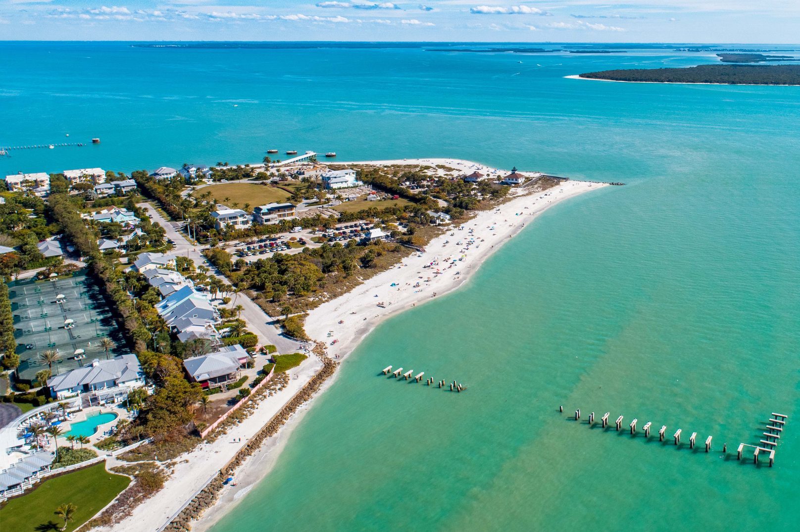 Beach curves along the coastline, dotted with people and umbrellas; nearby houses and trees are adjacent to the turquoise ocean.