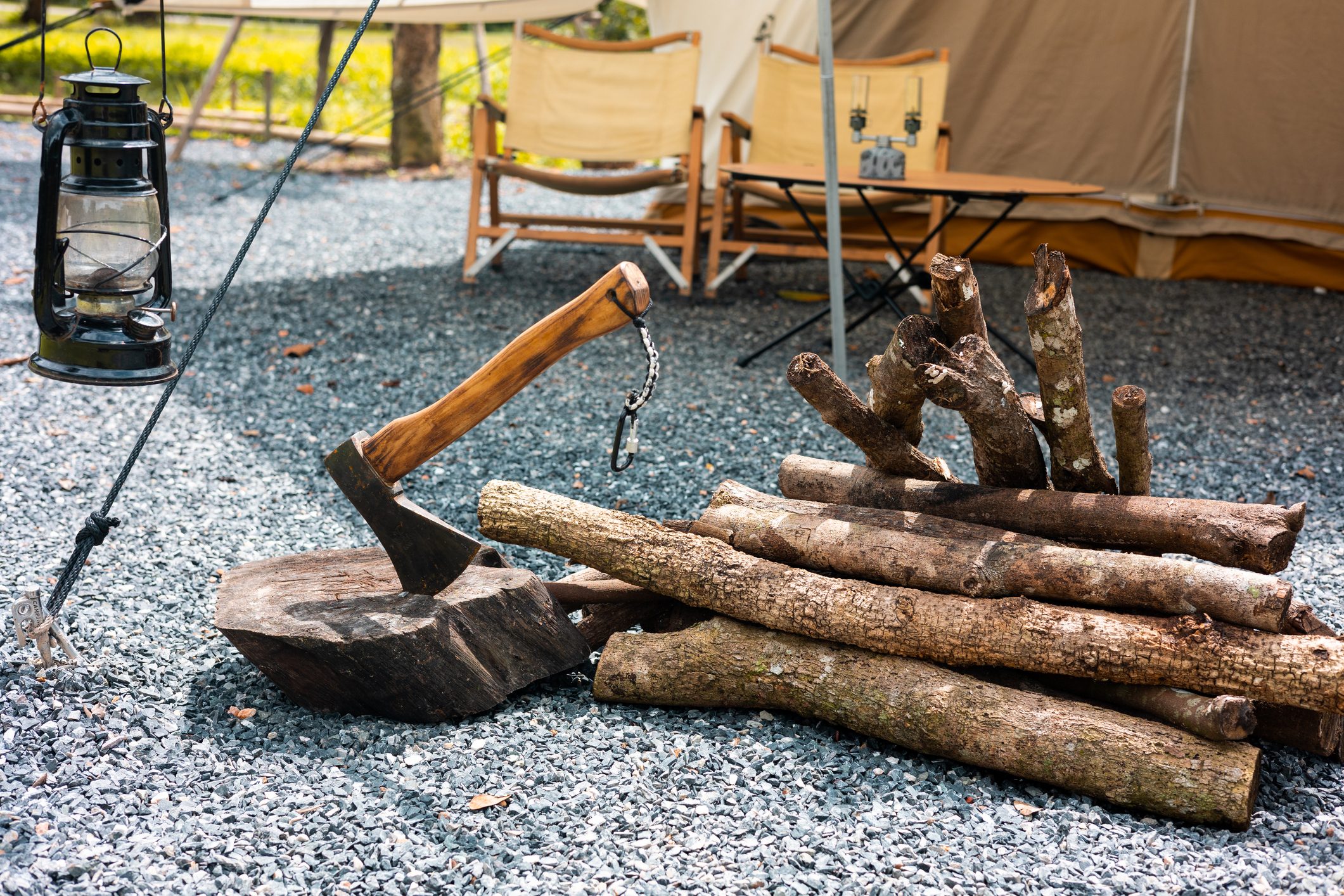 Axe embedded in wooden block; pile of logs nearby. Lantern hangs from rope. Camping chairs and tent visible in gravel-covered campsite.