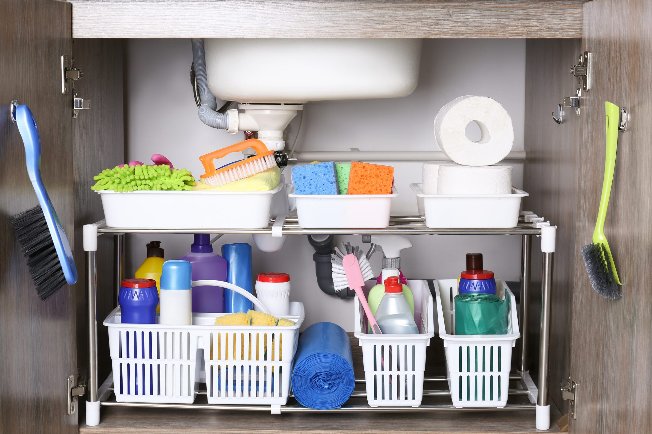 Cleaning supplies organized under a sink within a cupboard; includes brushes, sponges, bottles, and cloths, neatly stored in white baskets.