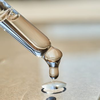 A glass dropper releases a liquid droplet onto a smooth, reflective surface in soft lighting.