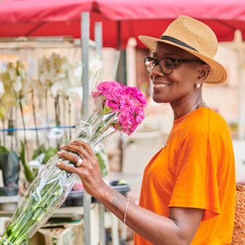 Person holding pink flowers, smiling, wearing a straw hat and orange shirt, standing near an outdoor flower market with plants and red umbrella.