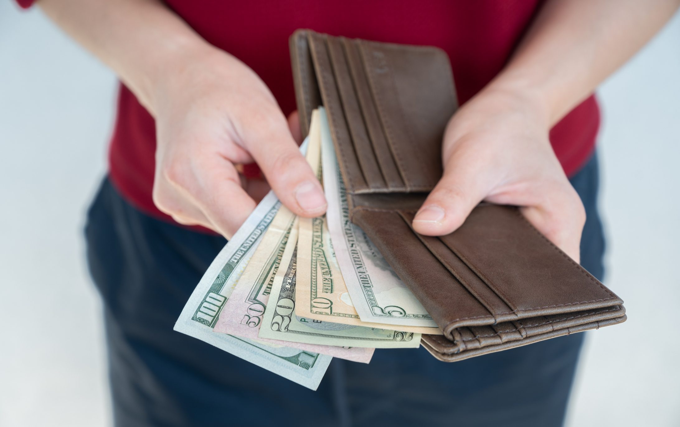 Hands hold an open brown wallet revealing multiple U.S. dollar bills, in an indoor setting.