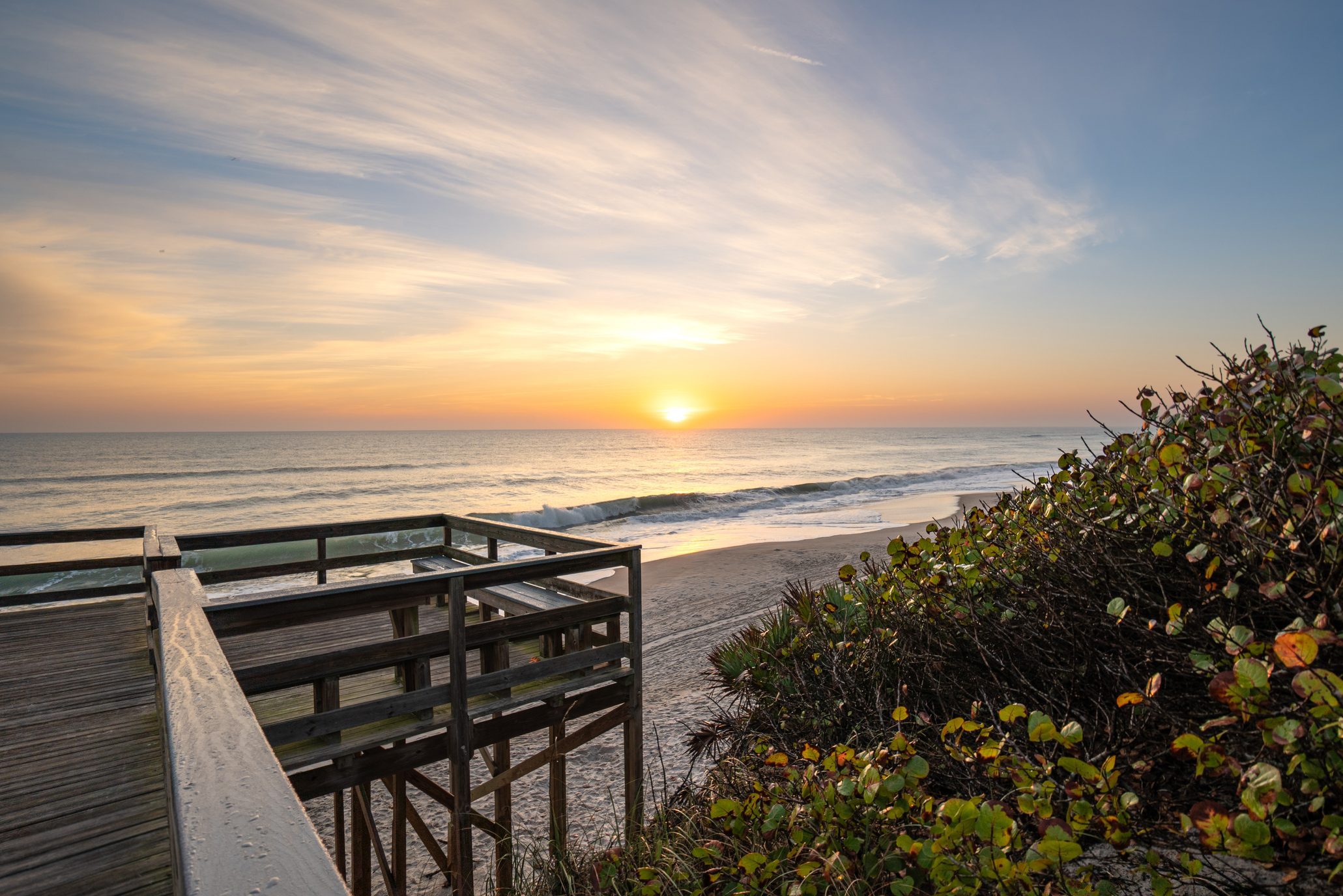 Wooden deck overlooks beach, capturing tranquil sunrise over the ocean. Bushes with green leaves frame the scene.