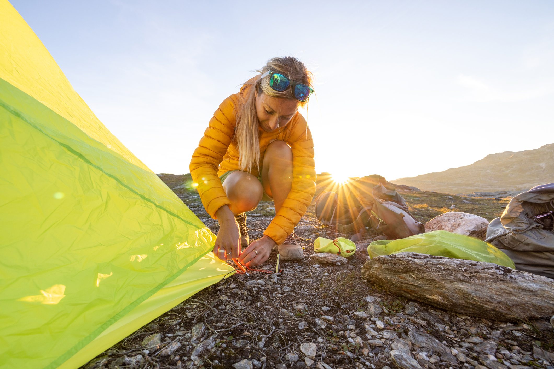 Person secures a tent on rocky ground, surrounded by backpacks, under a bright sun with mountains in the distance.