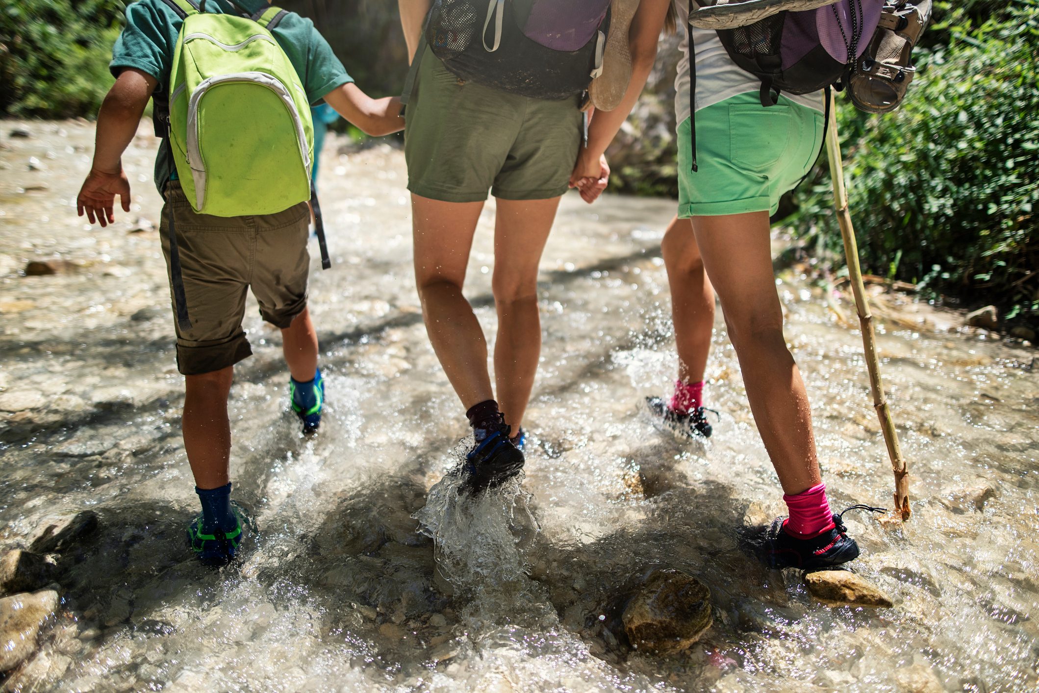 Three people wade through a shallow, rocky stream, holding hands. They're wearing shorts, backpacks, and walking shoes. Sunlight filters through surrounding greenery.