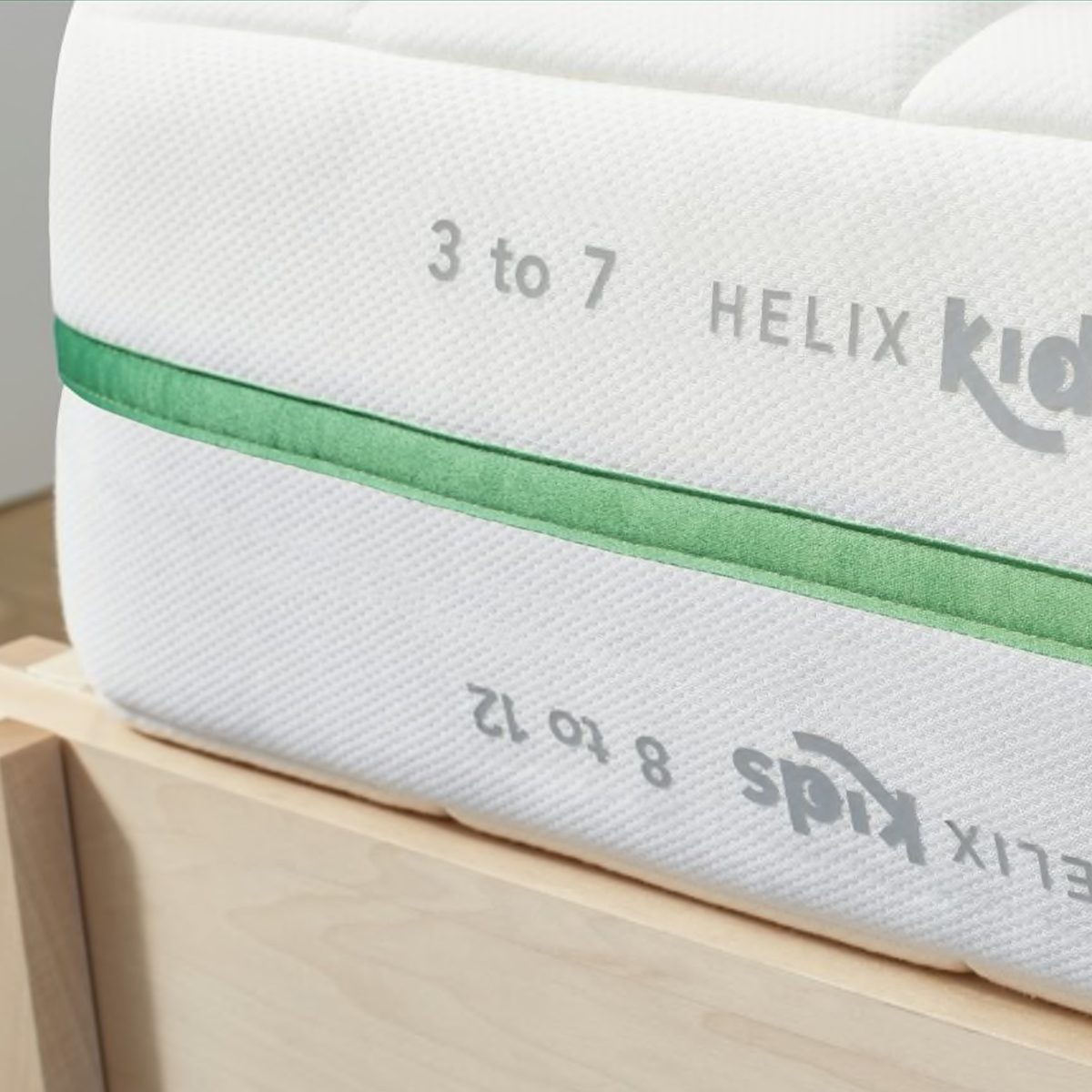 Mattress rests on a wooden frame, featuring a label "HELIX Kids, 3 to 7, 8 to 12" with a green stripe accent.