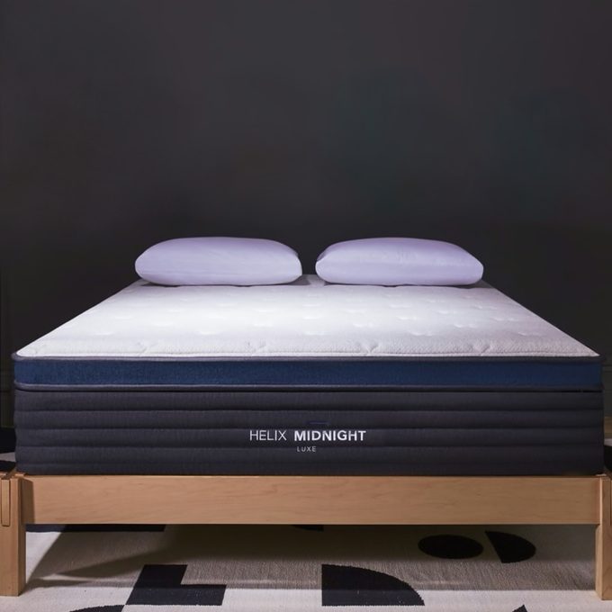 A bed with two pillows rests on a wooden frame against a dark background. The mattress label reads 