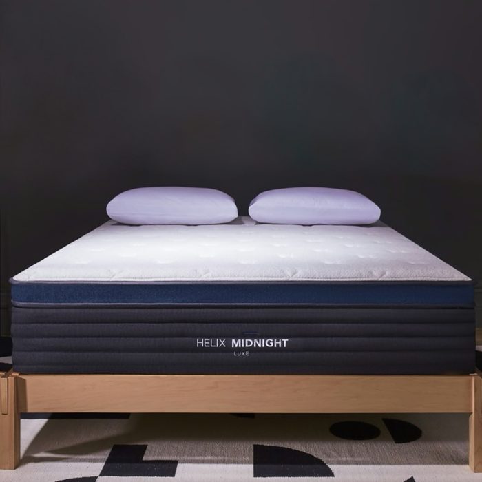 A bed with two pillows rests on a wooden frame against a dark background. The mattress label reads 