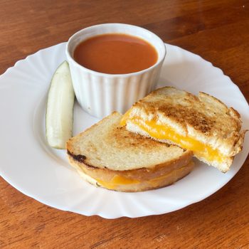 Grilled cheese sandwich sits on a white plate beside a pickle slice and small bowl of tomato soup on a wooden table.