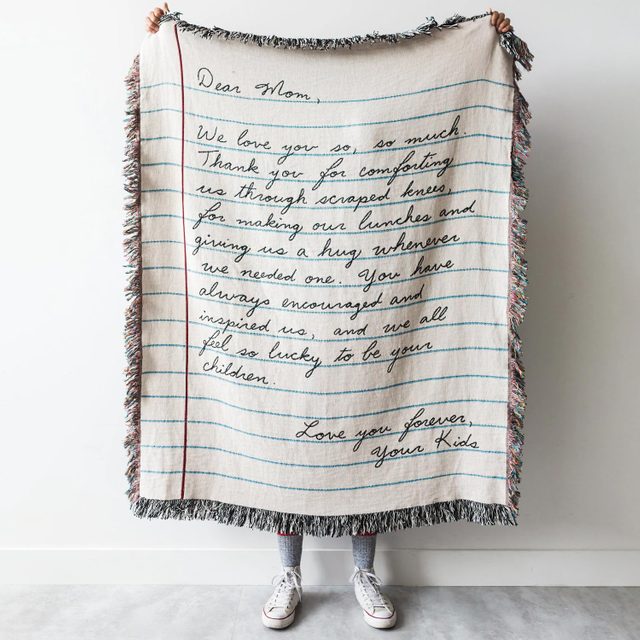 Fringed blanket displays handwritten letter expressing gratitude to mom, covering person from shoulders to feet, standing in a plain room.