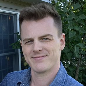 A person stands smiling, wearing a blue shirt, outdoors near a window and green leafy plants.