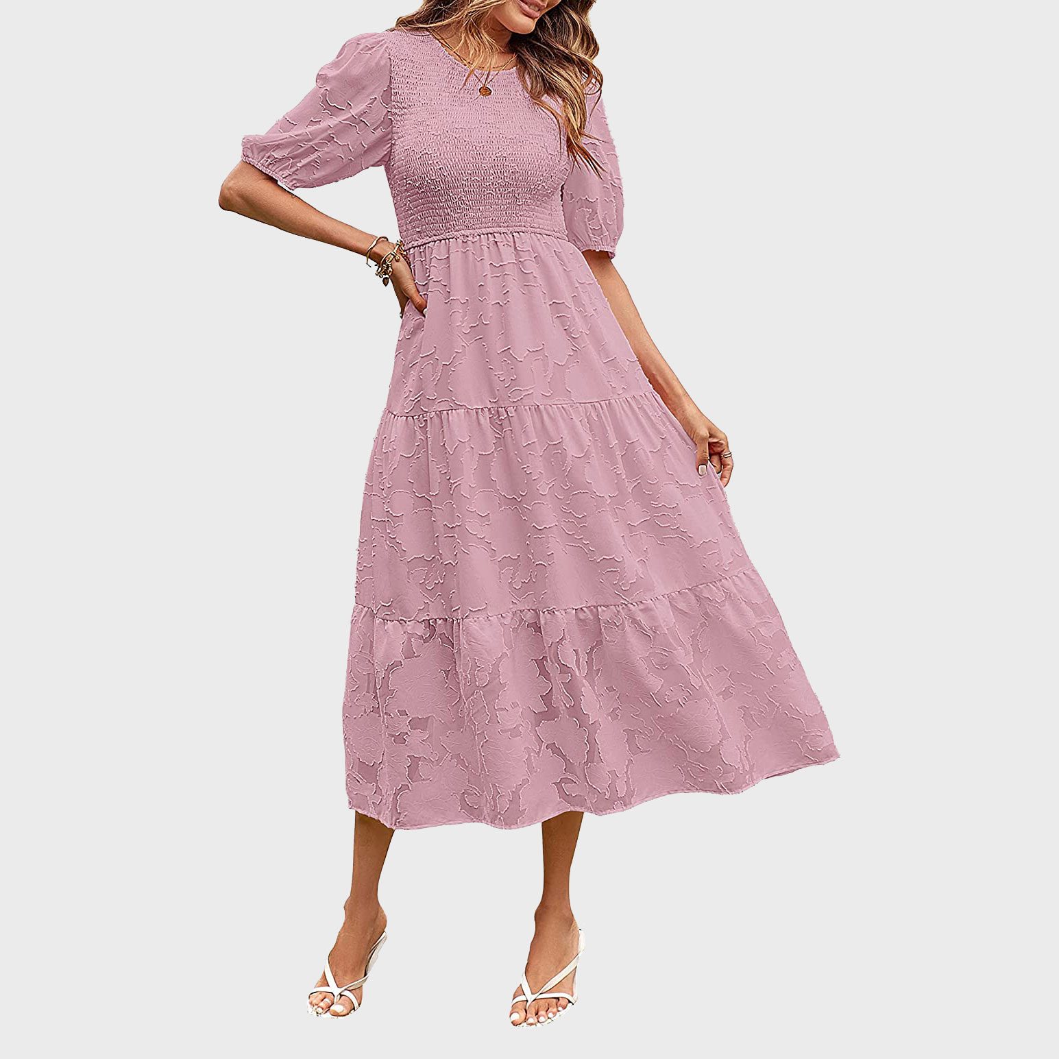 Woman wearing a light pink, textured maxi dress with puffed sleeves, standing against a plain white background, hands casually touching sides.