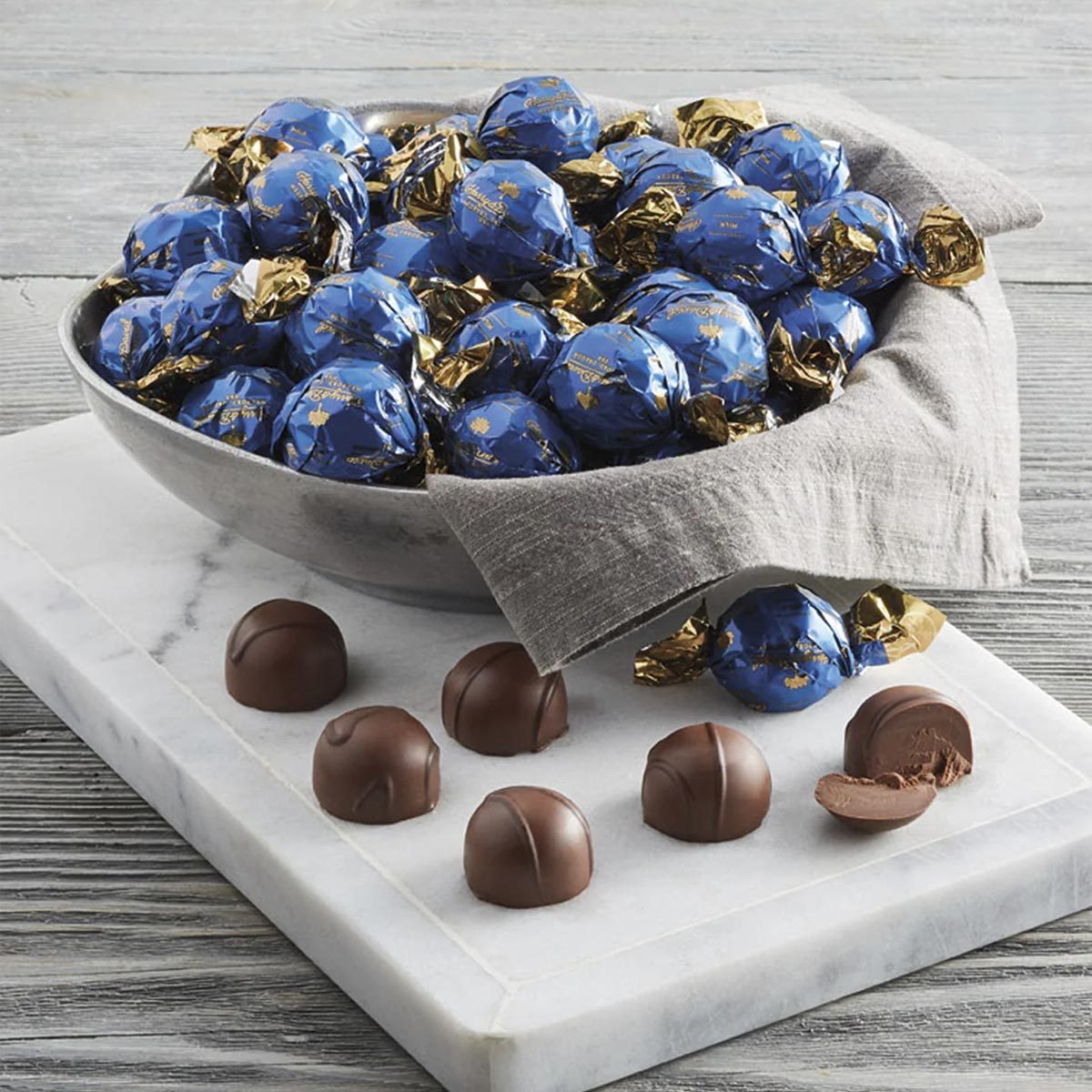 Bowl of blue-wrapped chocolates sits on a marble slab with unwrapped chocolates scattered around, set on a wooden table.