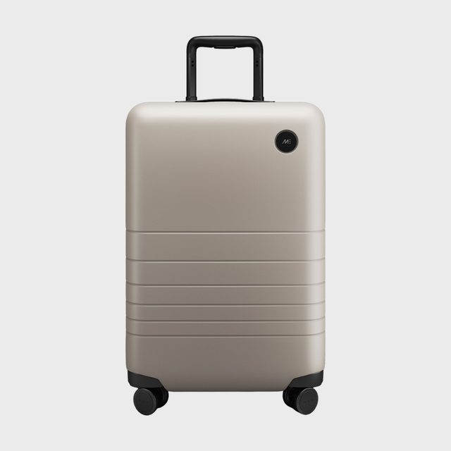 Suitcase stands upright with extended handle; smooth surface features horizontal grooves and four wheels, resting against a plain gray background.