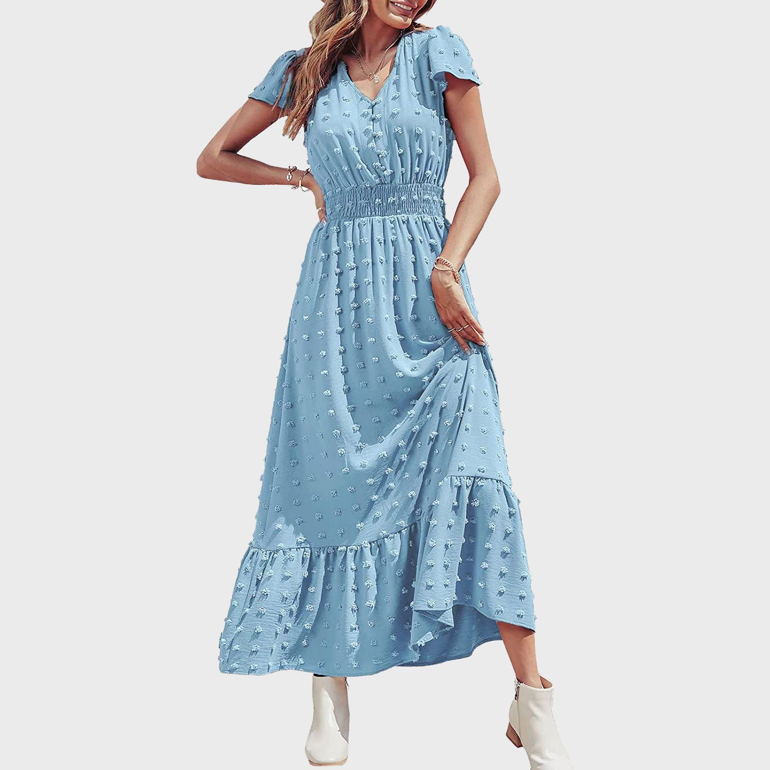A woman holds her light blue, textured maxi dress, smiling, posing in white ankle boots on a plain white background.